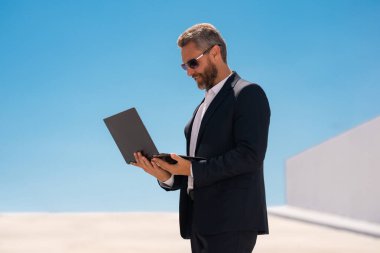 İş adamı dışarıda dizüstü bilgisayar kullanıyor. Olgun iş adamı dizüstü bilgisayar kullanıyor. Dizüstü bilgisayarı olan kendine güvenen bir iş adamı. İnternette iş. İş adamı laptoptaki verileri kontrol ediyor. Takım elbiseli iş adamı internette çalışıyor.