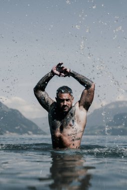 Seksi yaz erkeği. Suda ıslak kaslı erkek vücudu. Çıplak seksi erkek vücudu. İnsan yaz doğasında suya su sıçratır. Yaz plajında poz veren formda bir adam. Dağ gölünde su sıçratan güçlü bir adam. Yaz gezisi