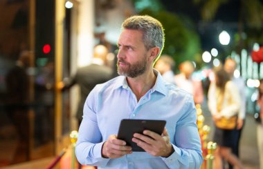 Gece sokakta tablet kullanan bir iş adamı. İş adamı dışarıda dijital tablet üzerinde çalışmayı planlıyor. Neon şehir ışıkları altında bir tablet üzerinde gezinen bir iş adamı. Serbest çalışan bir iş adamı. Gece işi.