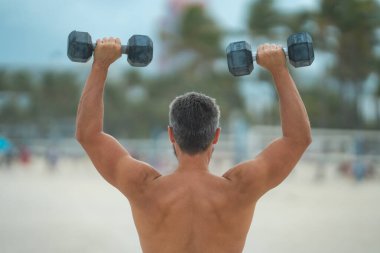 Spor adamının dambılları açık havada. Spor aktivitelerine katılan atletik bir adam. Formda adam geri döndü. Spor egzersizleri. Spor seansındaki üstsüz adam, geri döndü. Dambıllı açık hava eğitimi