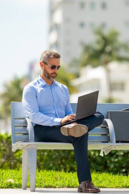 Kendine güvenen işadamı bankta dizüstü bilgisayarı ve parktaki telefonuyla oturuyor. Zengin iş adamı. Olgun iş adamı tatilin tadını çıkarıyor. İş adamı parktaki bankta mola veriyor. Yaz işleri.