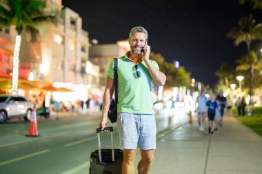Gece Miami sokaklarında telefon kullanan bir gezgin. Gezgin, yaz gezisi sırasında telefon etmiş. Gezgin telefonda konuşuyor. Gezgin telefonunu kontrol ediyor. Bir gece şehrinde bavuluyla bir turist.