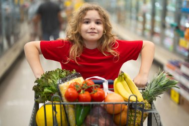 Süpermarkette el arabasıyla çocuk alışverişi. Çocuk markette taze sebze seçiyor. Yiyecek satan bir çocuk. Markette ürün seçen çocuk. Bakkalda taze yiyeceği olan çocuk