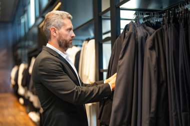 Bir adam butikte alışveriş yaparken klasik bir terzi kıyafeti giyer. Başarılı bir iş adamı için klasik bir kıyafet. Takım elbiseli modaya uygun bir mağaza. Olgun bir adam bir takım elbise seçer. Bir erkek resmi etkinlik için takım elbise seçer.