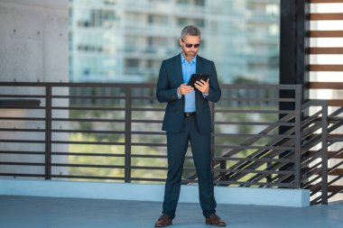 Açık havada tabletle başarılı bir iş adamı. Kendine güvenen iş adamı dışarıda tablet kullanıyor. Olgun iş adamı finans verilerini kontrol ediyor. Zengin iş adamı bankacı tabletteki sözleşmeyi gözden geçiriyor