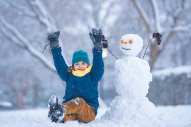 Soğuk bir kış gününde kış parkında bir çocuk. Kışın karla oynayan sevimli bir çocuk. Kardan adamla komik çocuk portresi. Noel 'de kış aktiviteleri. Karlı parkta kardan adam yapan çocuk