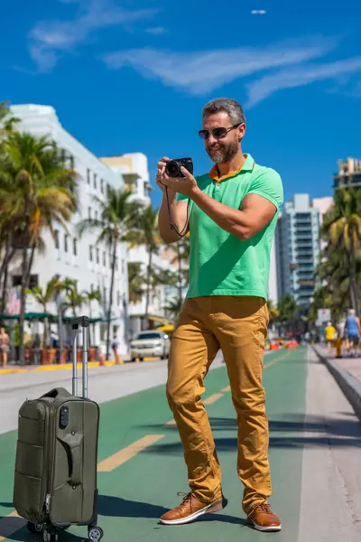 Miami sokaklarında fotoğraf çeken kameralı bir gezgin. Bir erkek gezgin, tropik bir şehirde yaz tatilinin tadını çıkarır. Yaz şehrinde bavullu bir turist. İnsan gezgin şehirde yürür