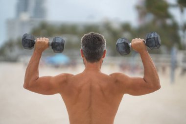 Spor adamının dambılları açık havada. Spor aktivitelerine katılan atletik bir adam. Formda adam geri döndü. Spor egzersizleri. Spor seansındaki üstsüz adam, geri döndü. Dambıllı açık hava eğitimi