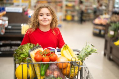 Süpermarkette el arabasıyla çocuk alışverişi. Çocuk markette taze sebze seçiyor. Süpermarkette yiyeceği olan bir çocuk. Süpermarkette ürün seçen bir çocuk. Süpermarkette taze yiyeceği olan çocuk