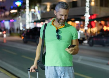 Man Turist gece Miami sokaklarında telefon kullanıyor. Yaz gezisi sırasında turist telefonları. Turist telefonda konuşur. Gezgin adam telefonu kontrol ediyor. Gece şehrinde bavullu bir turist.