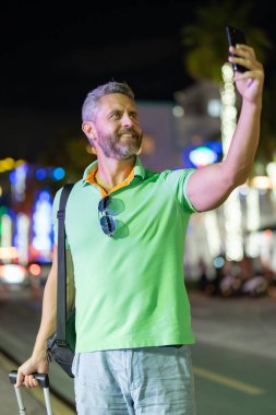 Hispanik gezgin gece Miami sokaklarında selfie çekiyor. Gece şehrinde telefonu olan bir gezgin. Yaz turistleri selfie çekiyor. Gezgin bavullu bir selfie için poz veriyor. Şehirde gezgin