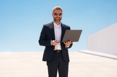 İş adamı dışarıda dizüstü bilgisayar kullanıyor. Dizüstü bilgisayarda çalışan başarılı bir adam. Dizüstü bilgisayarı olan kendinden emin bir iş adamı. İnternette iş. Şehir işadamı dışarıda dizüstü bilgisayardan verileri kontrol ediyor. Takım elbiseli adam internette çalışıyor.