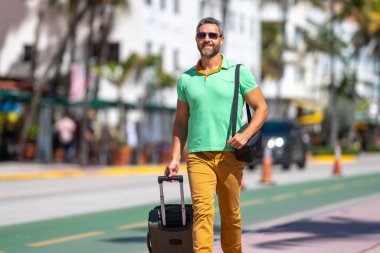 Tropikal şehirde bavul taşıyan bir turist. Yaz tatilinde bir erkek turist. Yaz şehrinde bavullu bir turist. Erkek turist şehirde yürüyor. Seyahat yaşam tarzı. Yaz tatili kaçamağı