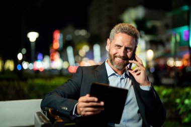 İş adamı telefonda konuşuyor ve tablet kullanıyor. Tabletli bir iş adamı. Tabletli ve telefonlu bir iş adamı. İş adamı tablet üzerinde çalışır, bankta oturur. Gece işi.