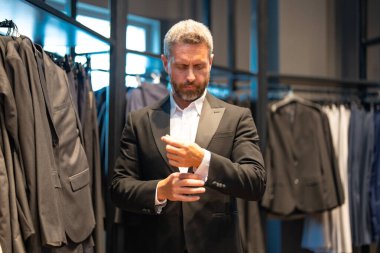 Olgun bir adam kıyafeti için mükemmel bir takım seçer. İyi giyimli bir erkek model bir butikte poz veriyor. Perakende satış asistanı şık bir adamın mükemmel takımı bulmasına yardım eder. Mağaza müdürü şık bir takım elbise ayarlar.