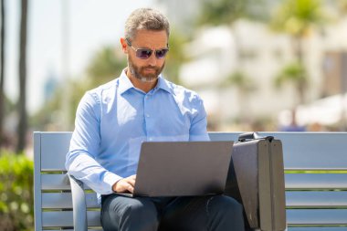 Kendine güvenen bir iş adamı bankta dizüstü bilgisayarı ve parktaki telefonuyla oturuyor. Başarılı bir iş adamı. Olgun iş adamı tatilin tadını çıkarıyor. İş adamı parktaki bankta mola veriyor. Yaz işleri.