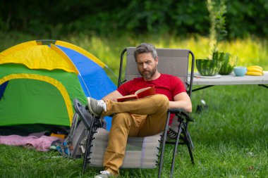 Kampta kitap okuyan bir adam. Doğaya yaz gezisi. Orman kampında dinlenen adam. Kamp gezisi. Yürüyüşten sonra çadırın yanında dinleniyor. Barışçıl bir kamp alanında özgürlük tatili. Piknikteki Adam