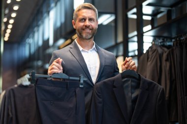 Olgun bir adam kıyafeti için mükemmel bir takım seçer. İyi giyimli bir erkek model bir butikte poz veriyor. Perakende satış asistanı şık bir adamın mükemmel takımı bulmasına yardım eder. Mağaza müdürü şık bir takım elbise ayarlar.