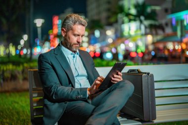 Dışarıda tablet üzerinde çalışan bir iş adamı. Tabletli bir iş adamı. Gece sokağında tabletli bir iş adamı. Tablet kullanan bir işadamı, bankta oturur. Gece işi.
