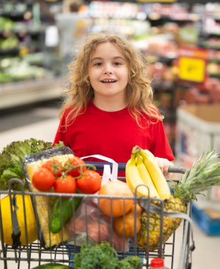 Süpermarkette el arabasıyla çocuk alışverişi. Çocuk markette taze sebze seçiyor. Süpermarkette yiyeceği olan bir çocuk. Süpermarkette ürün seçen bir çocuk. Süpermarkette taze yiyeceği olan çocuk