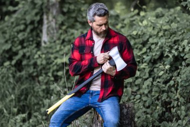 Ekose gömlekli oduncu ve kot pantolonlu. Yaz tahtasında büyük bir balta taşıyor. El işi. Doğal ortam. Ormanda baltası olan oduncu. Çalışkan adam. Baltası ya da baltası olan kaba sakallı bir adam.