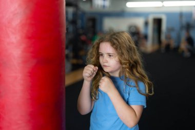 Boks torbasını yumruklayan çocuk. Beden eğitiminde genç boksör. Boks eldiveni giyen çocuk. Çocuk dövüş sanatları çalışıyor. Güçlü bir genç dövüşçü portresi. Sportif çocuk yumruğunu gösteriyor. Aktif çocuk egzersiz yapıyor