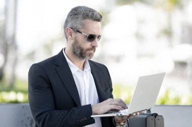 Kendine güvenen bir iş adamı bankta dizüstü bilgisayarı ve parktaki telefonuyla oturuyor. Başarılı bir iş adamı. Olgun iş adamı tatilin tadını çıkarıyor. İş adamı parktaki bankta mola veriyor. Yaz işleri.