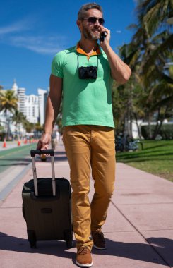 Miami sokaklarında kamera ve bavul taşıyan bir gezgin. Bir erkek gezgin, tropik bir şehirde yaz tatilinin tadını çıkarır. Yaz şehrinde bavullu bir turist. İnsan gezgin şehirde yürür