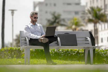 Kendine güvenen bir iş adamı bankta dizüstü bilgisayarı ve parktaki telefonuyla oturuyor. Zengin iş adamı. Olgun iş adamı tatilin tadını çıkarıyor. İş adamı parktaki bankta mola veriyor. Yaz işleri.