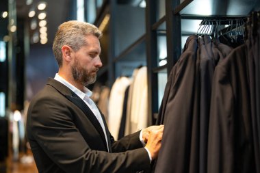 Takım elbiseli, butikte şık bir adam. Bir iş adamı mağazada şık bir takım elbise seçer. Modayı takip eden bir müşteri resmi kıyafet alırken ceketi inceler. Klasik kıyafetli çekici bir manken.