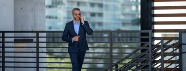 İş adamı telefonda konuşuyor ve yürüyor. Kendine güvenen bir iş adamı dışarıda telefonla konuşuyor. İş telefonu konuşması. Şirket lideri telefonda konuşmakla meşgul. Zengin iş adamı. İş çağrısı