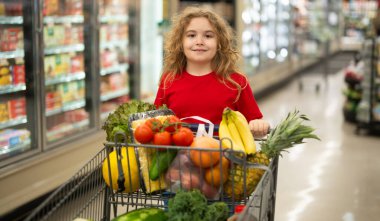 Süpermarkette el arabasıyla çocuk alışverişi. Çocuk markette taze sebze seçiyor. Yiyecek satan bir çocuk. Markette ürün seçen bir çocuk. Bakkalda taze yiyeceği olan çocuk