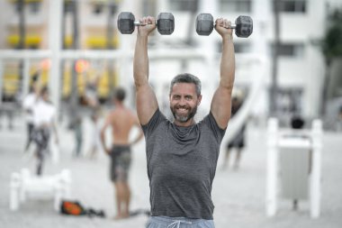 Açık havada dayanıklılık eğitimi. 40 'lı yaşlarda erkek spor yapıyor. Halterleri kaldıran yakışıklı vücut geliştirici. Fitness modeli eğitimi. Spor salonu açık havada. Sporcu adam spor yapıyor.