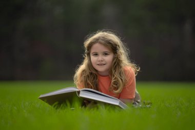 Çimlerin üzerinde kitap okuyan bir çocuk. Çocuk açık havada kitap okumaktan zevk alıyor. Güneşli bir günde çayırda kitapla ders çalışan bir çocuk. Doğada öğrenme. Öğrenci çocuk kitap okuyor. Çocuk açık havada öğreniyor