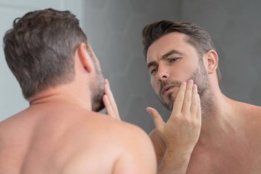 Mükemmel esmer adamın çene ve tenine dokunuşunun yakın plan portresi. Banyoda aynanın karşısında yüzüne dokunan yakışıklı bir adam. Mükemmel cilt. Erkek kozmetik, deri tedavisi. Hijyen ve cilt bakımı erkek yüzü