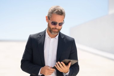 Açık havada tablet kullanan bir iş adamı. Tabletle başarılı bir yönetici. Şirket lideri tablet üzerinde çalışıyor. Zarif iş adamı tableti kontrol ediyor. Dijital aygıt kullanan işadamı