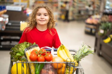 Süpermarkette el arabasıyla çocuk alışverişi. Çocuk markette taze sebze seçiyor. Süpermarkette yiyeceği olan bir çocuk. Süpermarkette ürün seçen bir çocuk. Süpermarkette taze yiyeceği olan çocuk