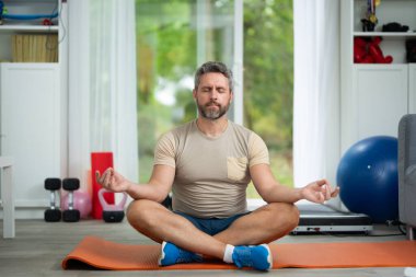 Yakışıklı orta yaşlı adam oturma odasında sabah yogası yapıyor. Formda bir adam, evde spor yaparken yoga minderinde geriniyor. Yoga pozları. Dairede sabah yoga egzersizi.