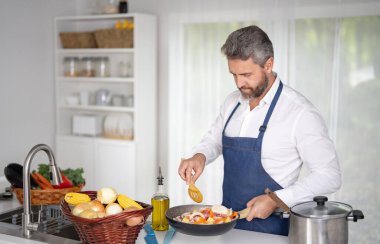 Adam mutfakta sebze doğruyor. Önlüklü aşçı modern mutfakta sağlıklı yemek hazırlar. Mutfakta sebzeli bir aşçı. 40 'lı yaşlar ev mutfağında sebzelerle kahvaltı hazırlar.