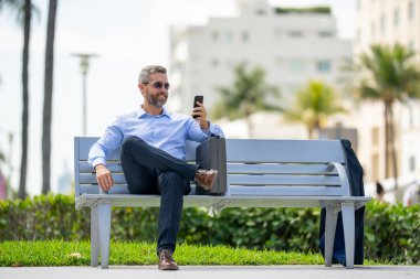 Kendine güvenen işadamı bankta dizüstü bilgisayarı ve parktaki telefonuyla oturuyor. Başarılı bir iş adamı. Olgun iş adamı tatilin tadını çıkarıyor. İş adamı parktaki bankta mola veriyor. Yaz işleri.