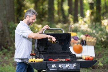 Şef lezzetli bir yemek için arka bahçedeki ızgarada et pişiriyor. Aktif bir adam yaz barbeküsü sırasında dışarıda yemek pişiriyor. Orta yaşlı İspanyol erkek ızgarada gurme yemekleri pişiriyor.