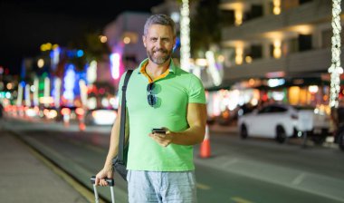Man Turist gece Miami sokaklarında telefon kullanıyor. Yaz gezisi sırasında turist telefonları. Turist telefonda konuşur. Gezgin adam telefonu kontrol ediyor. Gece şehrinde bavullu bir turist.