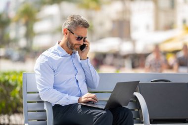 Kendine güvenen işadamı bankta dizüstü bilgisayarı ve parktaki telefonuyla oturuyor. Zengin iş adamı. Olgun iş adamı tatilin tadını çıkarıyor. İş adamı parktaki bankta mola veriyor. Yaz işleri.