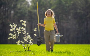 Bahçede ağaç yetiştiren bir çocuk. Çocuk kürekle toprak kazıyor ve ağaç dikiyor. Çocuk bahçıvan meyve ağaçları yetiştiriyor. Çocuklarla ağaç yetiştirmek. Çocuklar için bahçe işleri. Kürekli çocuk ağaç yetiştiriyor.