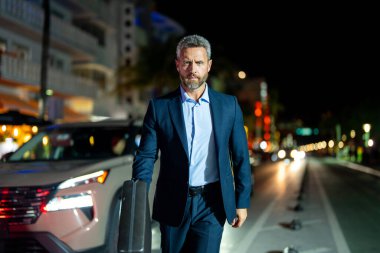 Gece vakti takım elbiseli bir iş adamı şehirde yürüyor. Takım elbiseli zengin bir adam. Lüks arabanın yanında bir iş adamı. Olgun bir iş adamı gece şehir ışıklarında yürür. İş stili. Zengin iş adamı
