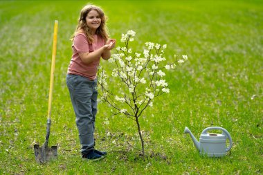 Çocuk bahçeye çiçek ve sulama ağacı. Bitkilerin bahar etkinliği. Çocuk kürek ve su bidonuyla çalışıyor. Çocuk meyve ağacı dikmiş. Bahçıvan çocuklar. Çocuk yetiştirme bitkisi ve ağaç bakımı