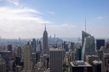 New York 'un panoramik manzarası ikonik Empire State binasını, modern gökdelenleri ve dağınık bulutlu açık mavi gökyüzünü gösteriyor..