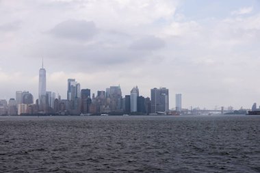 Yüksek gökdelenler ve bulutlu bir gökyüzünün yer aldığı, bir su kütlesi boyunca uzanan panoramik bir şehir manzarası. Görüntü, uzak mesafedeki modern mimari ve köprülerin karışımıyla şehir manzarasını yakalar..