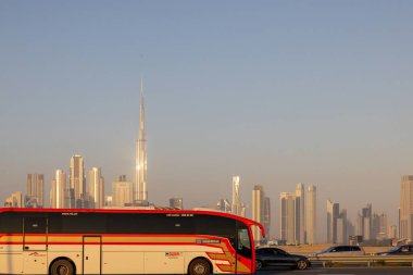 Dubai 'de modern gökdelenleri olan bir otoyolda kırmızı ve beyaz bir otobüs, açık mavi gökyüzünün altındaki ikonik Burj Khalifa da dahil..