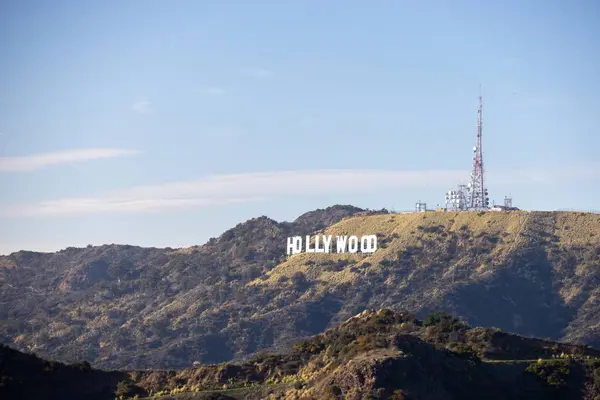 Yeşil dağlar ve açık mavi gökyüzü ile çevrili bir yamaçtaki ikonik Hollywood tabelası. Yakınlarda bir iletişim kulesi görülebiliyor ve manzaraya ekleniyor..